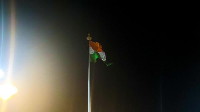 National tricolor flag of India hoisted on tall flagpole at night with street lighting.