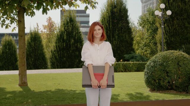 A redhead woman stands outside in a green area. She smiles at the camera while holding a laptop. Tall buildings are visible in the background under clear skies.