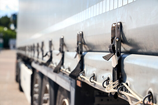 Close-up shot of heavy duty ratchet straps, hooks, tensioning buckles, side curtain fasteners securing grey tarp cover on commercial semi trailer cargo truck parked outdoors near highway