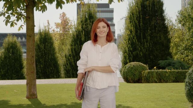 A woman with red hair turns to face the camera while holding a laptop in a green park. Trees and a fountain are visible in the background during daylight.