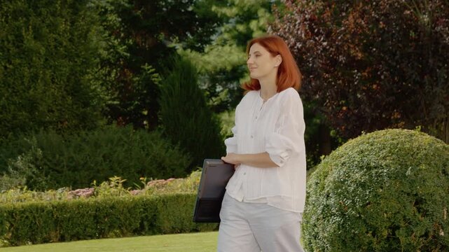 A female student walks through a park holding a laptop in her arms. She enjoys the day and the quiet surroundings as she moves along the path.