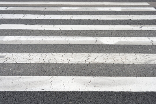 A close up shot of a pedestrian crosswalk with white stripes painted on weathered and cracked black asphalt pavement.