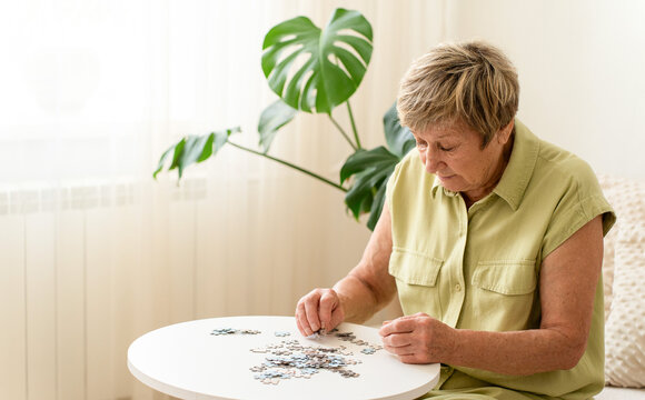 Senior woman actively engages her mind by putting together a puzzle on small table. Slow, mindful living, analog hobby, relaxing activity at home