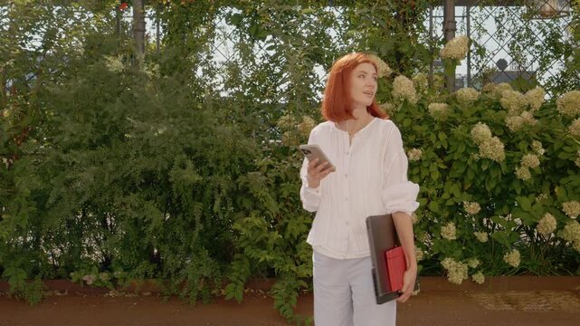 A female student stands on a campus path holding a laptop and her phone. She appears lost and is searching for the right way to go during the day.