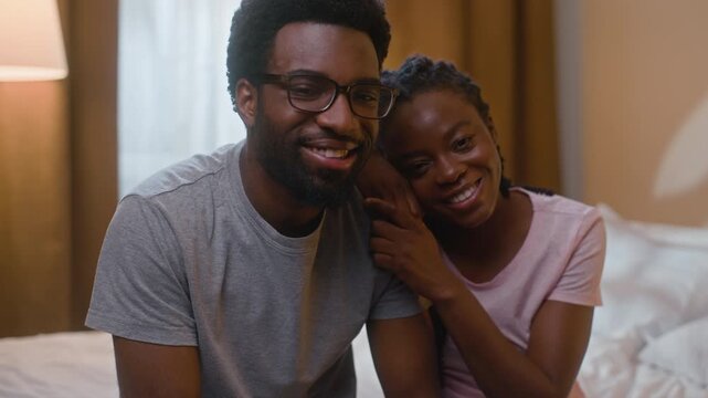 Happy African American married couple family hugging embracing sitting on bed looking at camera loving pair spouses portrait husband wife joyful relationship at home illuminated bedroom evening night