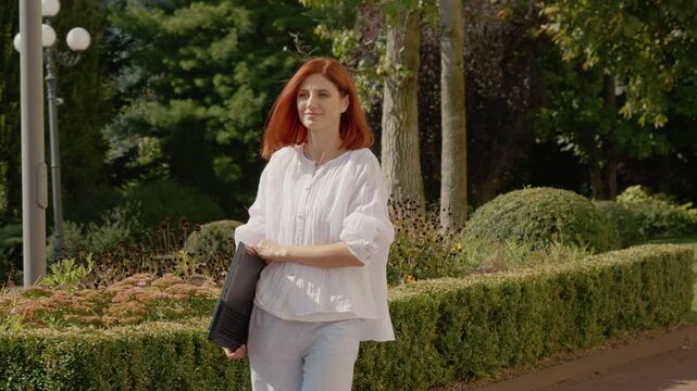 A female student is walking towards the university while holding a laptop. The campus is filled with green plants and trees.