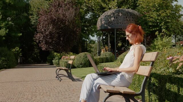 A student sits on a park bench in a green area. They focus on typing on a laptop, enjoying the warm weather in the park in the afternoon light.