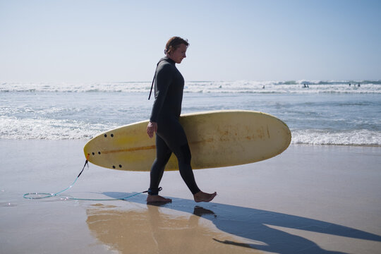  woman surfer in black wetsuit walking along sandy beach holding surfboard