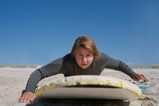  woman surfer in wetsuit practicing paddling and pop-up on surfboard at sandy beach
