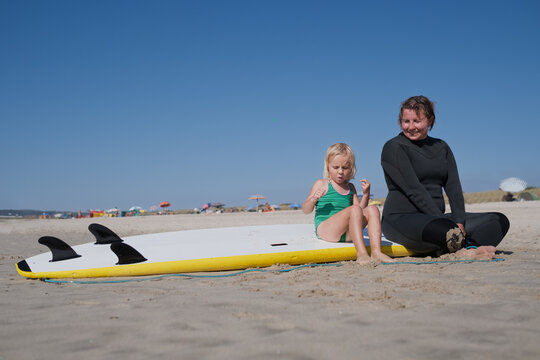  mother in wetsuit sitting on sand with little daughter on surfboard at beach