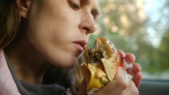 Closeup young woman sitting in car and hungrily eating delicious cheeseburger, taking large bites and enjoying fast food meal during quick lunch break on go. Woman eating juicy cheeseburger in car