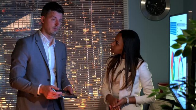 Two mixed race economic experts discuss causes of the world&rsquo;s economic crisis in a nighttime office with city skyscrapers visible through large windows.