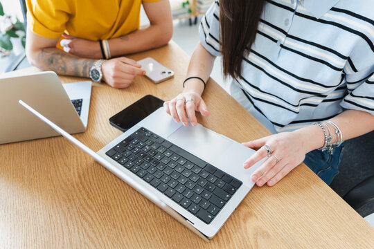 Two young colleagues sit together at a wooden table in an office. A woman types on a laptop keyboard while a man works on his own computer nearby during a project meeting.