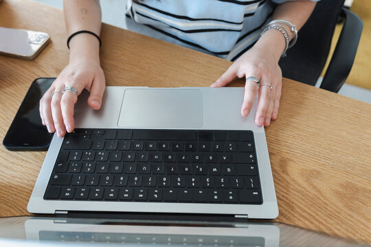 A young woman in a striped shirt sits at a wooden desk, actively typing on a silver laptop. Two smartphones rest nearby as she works on her daily professional tasks.