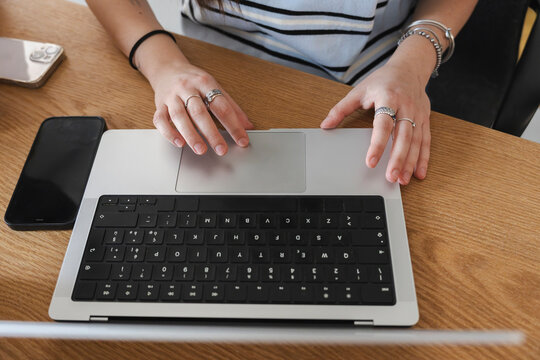 A woman working remotely at a wooden desk uses the trackpad on her laptop. A smartphone rests nearby while she navigates digital documents during the workday.