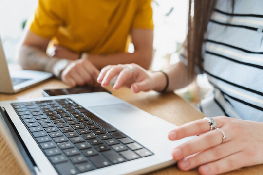 Two young colleagues are sitting together at a wooden table, working on their laptops in a bright office space. They are typing and collaborating on a modern digital project.