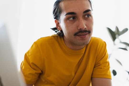 A young man with dark hair and a mustache wearing a yellow t-shirt sits indoors, looking thoughtfully toward a computer screen while working remotely from his home office.