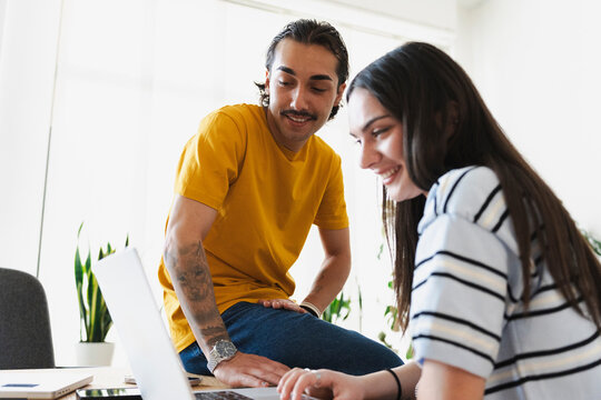 A young man and woman collaborate together in a bright modern office space. They look at a laptop computer screen and smile while working on a shared creative project.