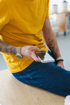 A tattooed man wearing a yellow t-shirt and jeans sits indoors and holds a modern smartphone in his hands, checking messages or browsing the internet during a casual workday.