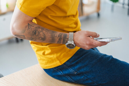 A man with a wolf tattoo on his arm wears a yellow t-shirt and blue jeans. He sits indoors while holding a modern smartphone and using the touchscreen to browse the internet.