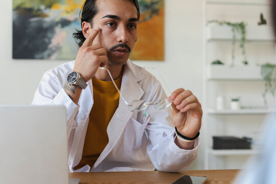 A male doctor in a white coat sits at his office desk, holding glasses and pointing to his eye while talking with a patient during a medical consultation.