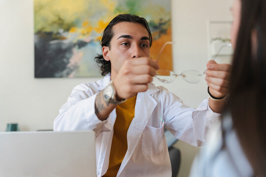 A young male optometrist wearing a white lab coat carefully holds up a pair of eyeglasses to show a patient during an eye examination inside a bright office.