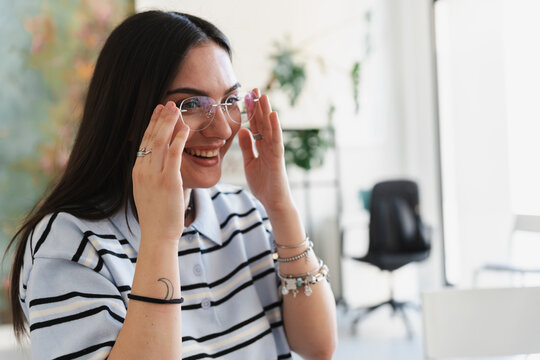 A young woman with long dark hair wearing a striped polo shirt smiles happily while putting on her new clear eyeglasses inside a bright modern office space.