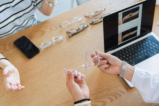 An optometrist hands a pair of new prescription eyeglasses to a customer during a consultation in a modern optical shop. A laptop with eye scans sits on the wooden table.