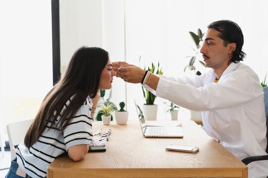 An optician in a white coat adjusts new eyeglasses on a young female customer sitting at a wooden desk inside a bright optical clinic during a professional consultation.