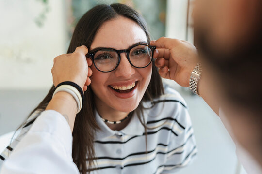 A cheerful young woman smiles brightly while trying on a new pair of stylish eyeglasses with the assistance of an optometrist in a modern optical shop.
