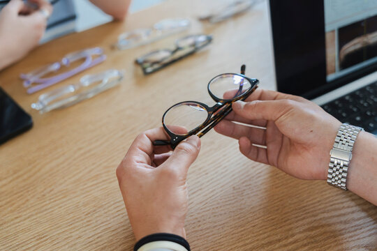 A person sits at a wooden table and holds a pair of stylish tortoiseshell eyeglasses for examination. Several other frames and a laptop are arranged on the desk surface.