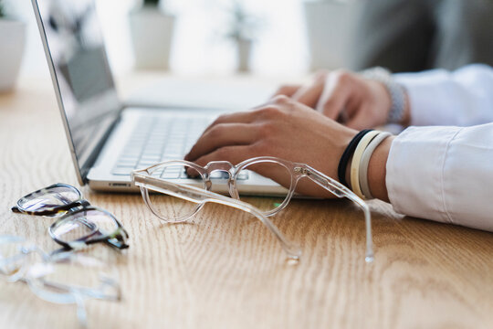 A professional person wearing a white shirt sits at a light wood desk and types on a laptop computer while several modern eyeglasses frames rest nearby.