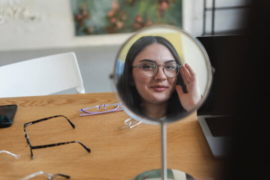 A young woman sits at a wooden desk and looks into a round mirror. She is trying on a pair of stylish eyeglasses, surrounded by several other frames scattered around her desk.
