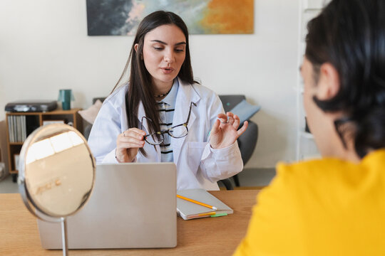 A female doctor in a white coat sits at a desk holding glasses while consulting a male patient in a bright office. They discuss vision correction options during an eye exam.