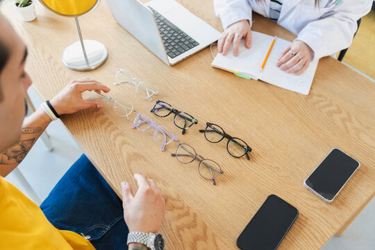 An optician and a customer sit at a wooden table in an optical shop. The man looks at several pairs of eyeglasses laid out in front of him to choose a new frame.