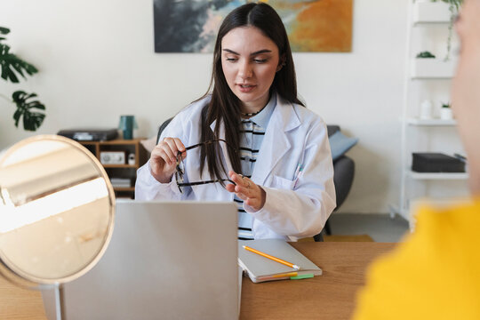 A female doctor wearing a white coat sits at a wooden desk holding a pair of eyeglasses, consulting with a patient during a medical appointment in a bright office.