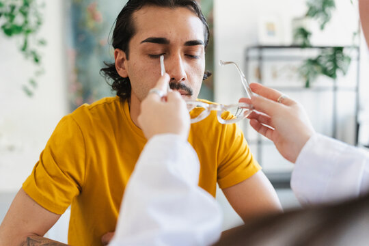 A young man with a mustache sits with his eyes closed while an optometrist adjusts and fits a new pair of clear-framed eyeglasses on his face during an eye exam.