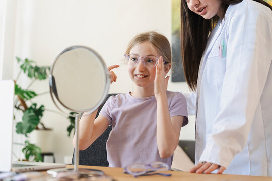 A young girl tries on new eyeglasses and looks in a mirror during an eye exam. A female optometrist stands next to her, assisting with frame selection in a modern clinic.