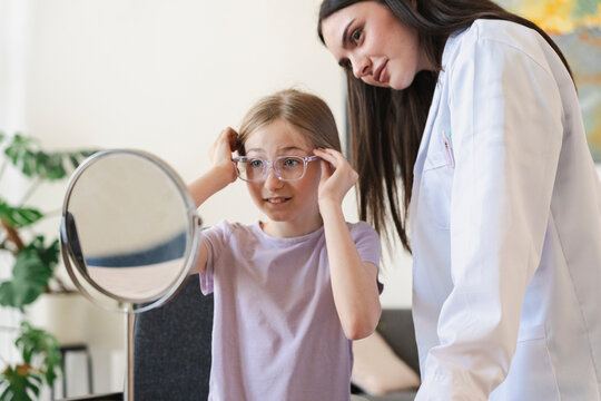An optometrist helps a young girl try on new eyeglasses while looking in a mirror. The female doctor smiles as the child tests the frames in a bright clinical office setting.