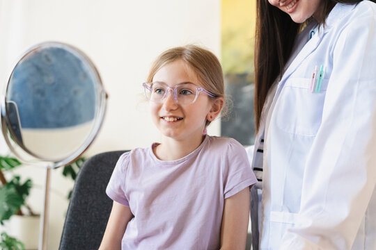 A young blonde girl wearing new prescription eyeglasses smiles happily during her eye exam while standing next to a friendly female doctor inside a bright medical clinic.