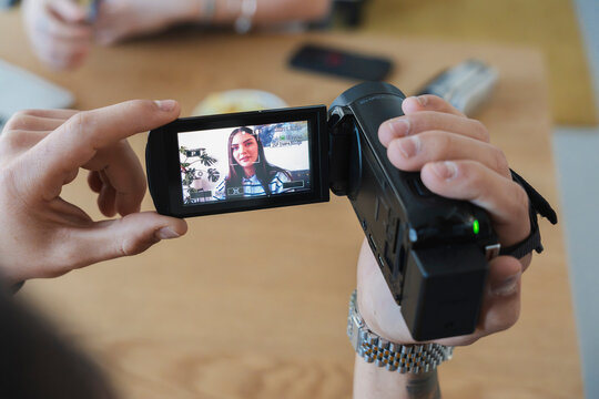 A person holds a black digital camcorder with both hands. They are viewing a young woman's face on the bright screen while recording a video clip indoors.