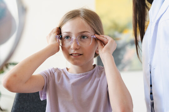 A young girl tries on new eyeglasses during an eye exam at a modern clinic. She carefully adjusts the stylish frames on her face while a professional doctor watches.
