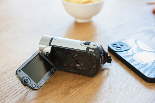 A silver digital camcorder with an open screen rests on a wooden table next to a smartphone. A blurred bowl sits in the background during a quiet indoor setting.