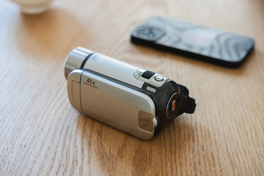 A silver digital video camera rests on a wooden table next to a smartphone. This setup highlights the contrast between traditional recording technology and modern devices.