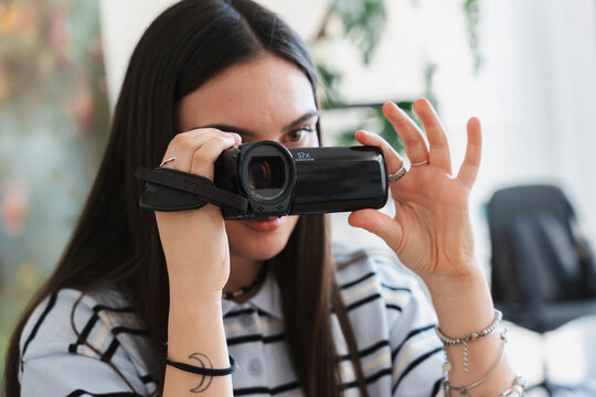 A young woman with long dark hair holds a small black digital video camera up to her eye, recording footage indoors. She wears a striped shirt and bracelets.
