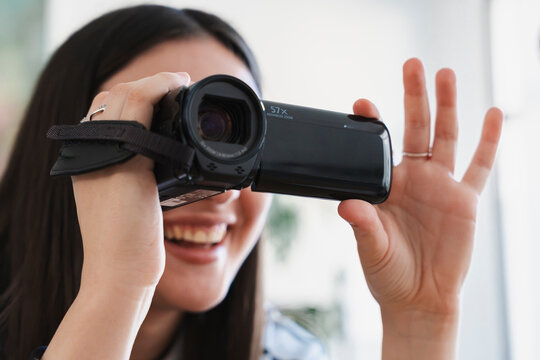 A smiling young woman with long dark hair holds a black digital camcorder up to record a video indoors, looking directly through the lens with a joyful expression.