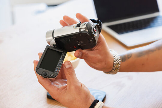 A person is holding a small digital camcorder in their hands, opening its display screen to check the device. They are sitting at a wooden desk with a laptop computer.