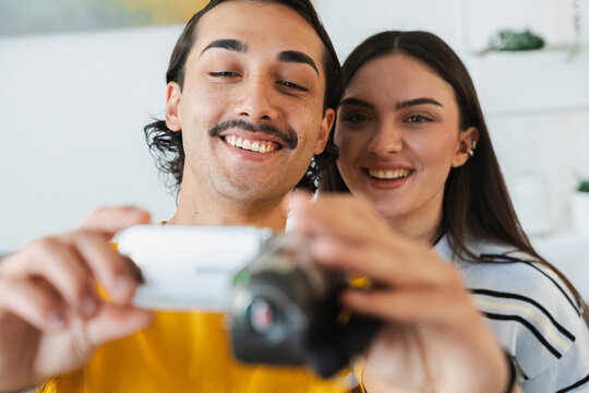 A smiling young man and woman stand together indoors while recording a video. They happily hold up a compact digital camera to capture a special moment at home.