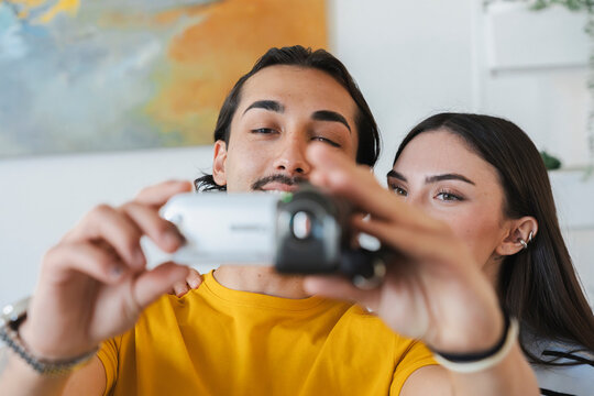A young couple smiling together at home while holding up a small digital video camera to record a video clip or vlog. They stand close together in a bright living room.