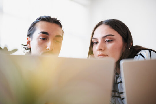 A young man and woman are focused on their laptops, collaborating on a project in a bright workspace. They study the screens together with concentrated expressions.